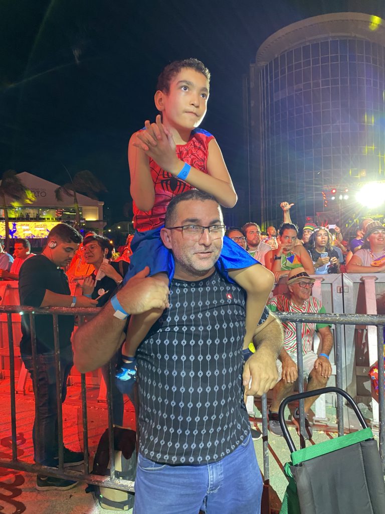 Fotografia tem Edgar que um homem branco, cabelo curto, usa óculos, está de blusa preta e calça jeans e nos seus ombros está seu filho João Victor, que criança com deficiência, vestida de homem aranha.  Atrás dele tem público geral do Marco Zero. 