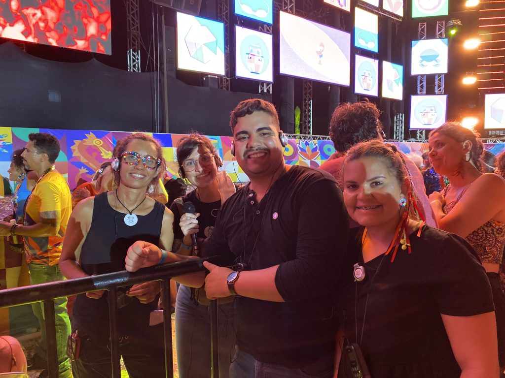 fotográfia contém quatro pessoas (três mulheres e um homem) todas de blusa preta e calcça jenas, com fones de ouvido em frente ao palco do Marco Zero.