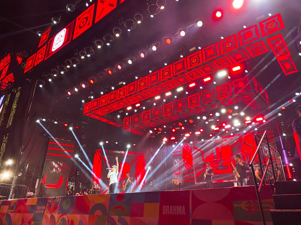 Fotografia no palco do marco zero todo iluminado de vermelho, ao centro tem cantor João gomes cantando com seus músicos. 