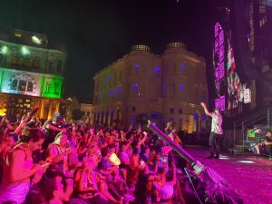 Um grupo de 25 pessoas com deficiência está reunido em frente ao palco. Atrás delas, uma plateia animada, com foliões em trajes carnavalescos. Ao fundo, edifícios históricos iluminados em tons de verde, azul e roxo compõem o cenário.