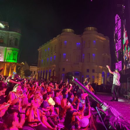 Um grupo de 25 pessoas com deficiência está reunido em frente ao palco. Atrás delas, uma plateia animada, com foliões em trajes carnavalescos. Ao fundo, edifícios históricos iluminados em tons de verde, azul e roxo compõem o cenário.