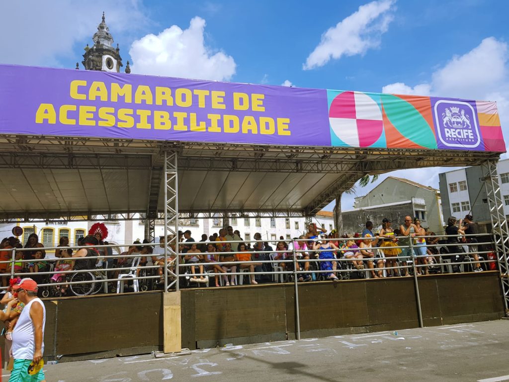 Fotografia com a estutura do Camarote da acessibilidade, com várias pessoas com deficiência esperando o desfile do Galo da Madrugada. Atrás do Camarote tem Igreja do Carmo e céu azul com poucas nuvens. 