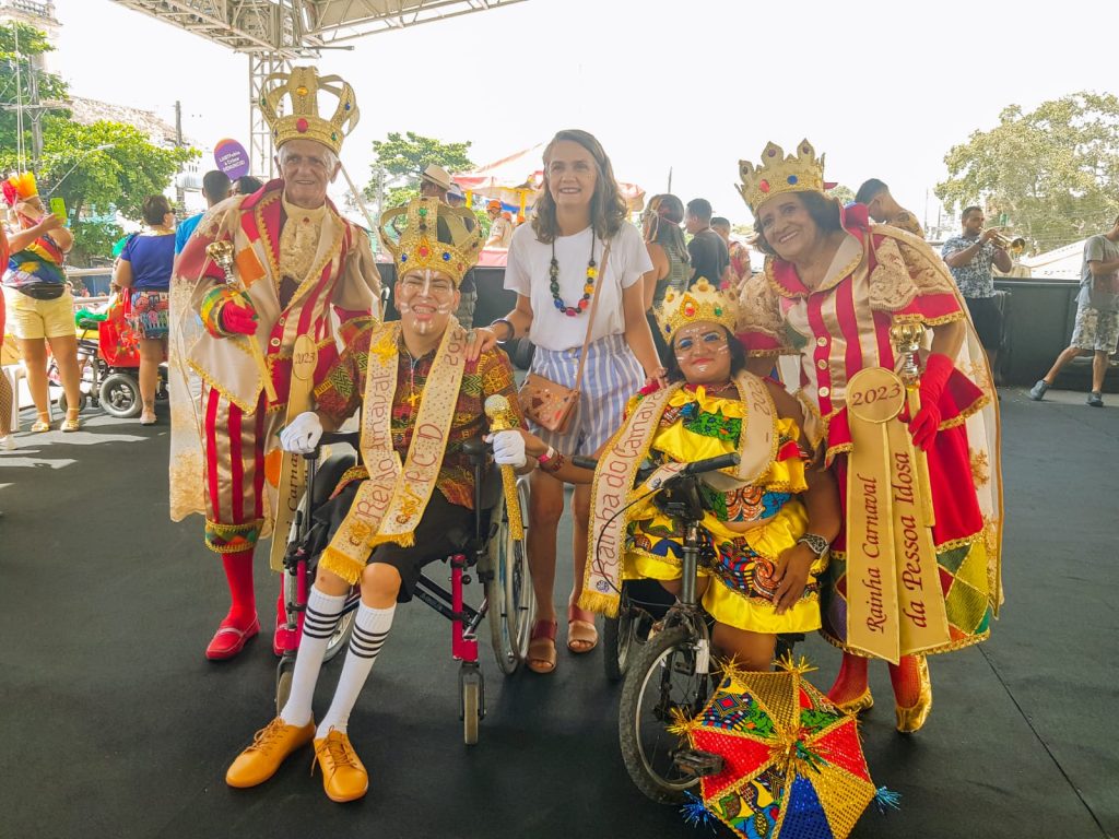 Fotografia com o Rei e rainha da pessoa com deficiência na cadeiras de rodas e ao lado deles tem o Rei e Rainha da pessoa idosa e no meio está Ana Rita Suassuna que é secretária de Desenvolvimento Social, Direitos Humanos, Juventude e Políticas sobre Drogas.