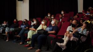 fotografia de uma sala de cinema com cadeiras vermelhas e várias pessoas espalhadas olhando para frente,