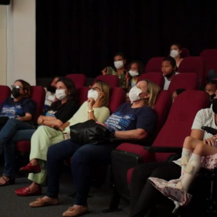 fotografia de uma sala de cinema com cadeiras vermelhas e várias pessoas espalhadas olhando para frente,