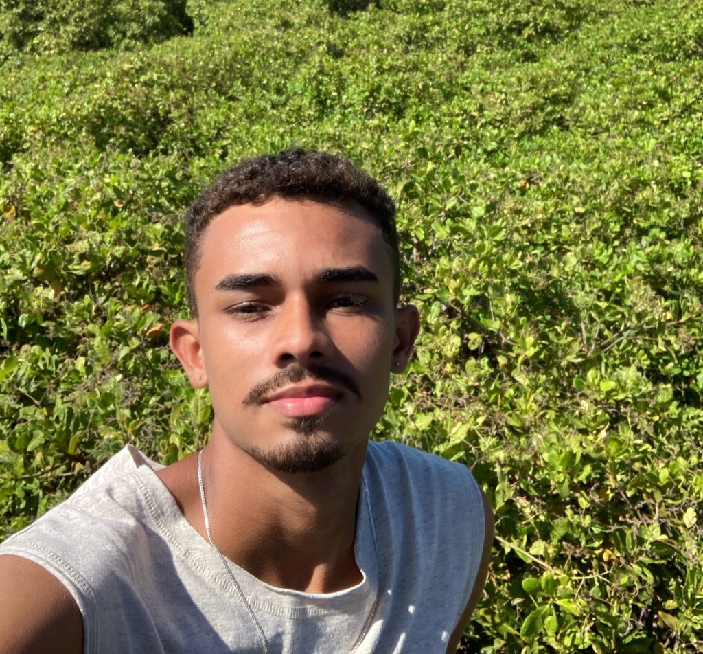 homem negro, cabelo curtos castanhos, bigode, usando regata cinza. Ao fundo várias plantas.