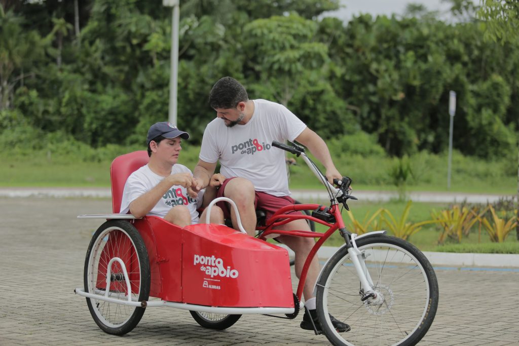 Fotografia: Um homem branco, com cabelos curtos e pretos, usa barba, está vestindo uma camisa branca com a frase "Ponto de Apoio" em cinza e vermelho. Ele está sentado em uma bicicleta vermelha e branca. Ao seu lado, há um rapaz branco com cabelos curtos e pretos, usando um boné azul e sentado em um carrinho vermelho com a frase "Ponto de Apoio" em branco. Ao fundo, há várias árvores e vegetação.