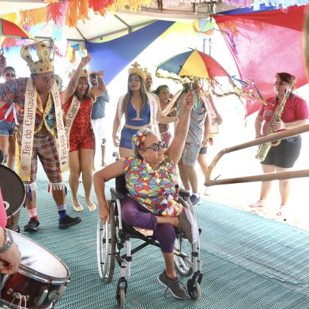 Fotografia de uma mulher parda, de cabelos grisalhos e óculos, sentada em uma cadeira de rodas enquanto segura uma sombrinha de frevo. Ao fundo, Luzia Santos, Rainha do Carnaval 2024, e Roberto Cabral, Rei do Carnaval 2024, acompanhados por outras pessoas fantasiadas e uma orquestra,