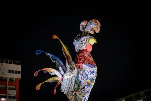Galo da Madrugada iluminado à noite, decorado com mosaicos coloridos, rendas e detalhes vibrantes. Seu bico amarelo e a cauda exibe penas estilizadas em tons de azul, verde, vermelho e amarelo. No fundo, ceú todo preto.