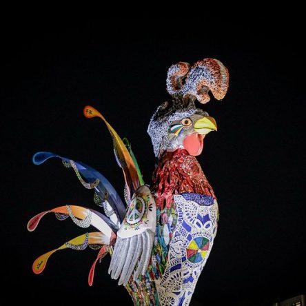 Galo da Madrugada iluminado à noite, decorado com mosaicos coloridos, rendas e detalhes vibrantes. Seu bico amarelo e a cauda exibe penas estilizadas em tons de azul, verde, vermelho e amarelo. No fundo, ceú todo preto.