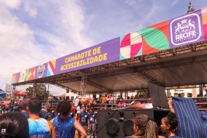 A imagem mostra o Camarote da Acessibilidade do Carnaval do Recife. O camarote é uma estrutura ampla e coberta, com uma grande faixa roxa na parte superior tem escrito "Camarote de Acessibilidade" em letras amarelas. Ao lado, há o brasão da Prefeitura do Recife e elementos gráficos coloridos. Na frente tem várias pessoas.