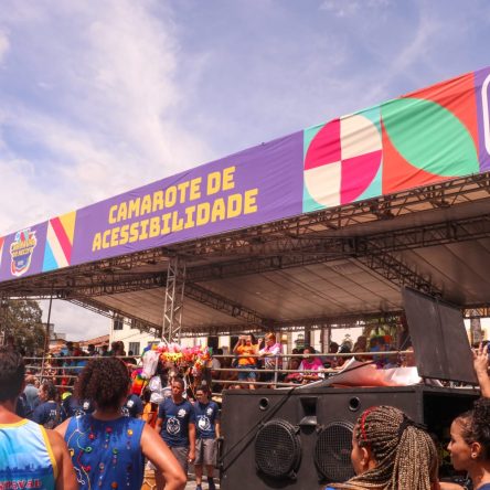 A imagem mostra o Camarote da Acessibilidade do Carnaval do Recife. O camarote é uma estrutura ampla e coberta, com uma grande faixa roxa na parte superior tem escrito "Camarote de Acessibilidade" em letras amarelas. Ao lado, há o brasão da Prefeitura do Recife e elementos gráficos coloridos. Na frente tem várias pessoas.