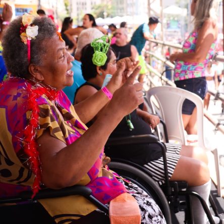 Fotografia de uma mulher parda, sorridente, vestindo roupas coloridas, sentada em uma cadeira de rodas de frente para uma grade. Ao fundo, várias pessoas trajando roupas carnavalescas estão sentadas.