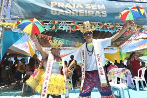 Na fotografia, Edilene, uma mulher parda, usa uma coroa dourada. Ela veste uma faixa branca no pescoço que tem escrito "Rainha do Carnaval", um body prateado, uma saia amarela e segura uma sombrinha de frevo. Ao seu lado, Ivan, um homem pardo, também usa uma coroa dourada com pedras coloridas, óculos escuros e colares pretos. Ele veste uma faixa branca no pescoço que tem escrito "Rei do Carnaval", uma camisa branca e calça xadrez, segura uma sombrinha de frevo. Ao fundo, há uma tenda com letreiro do projeto "Praia sem Barreiras".