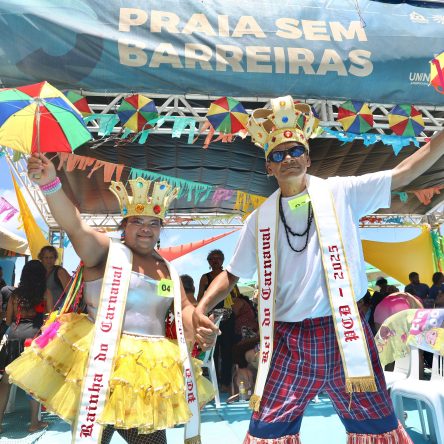 Na fotografia, Edilene, uma mulher parda, usa uma coroa dourada. Ela veste uma faixa branca no pescoço que tem escrito "Rainha do Carnaval", um body prateado, uma saia amarela e segura uma sombrinha de frevo. Ao seu lado, Ivan, um homem pardo, também usa uma coroa dourada com pedras coloridas, óculos escuros e colares pretos. Ele veste uma faixa branca no pescoço que tem escrito "Rei do Carnaval", uma camisa branca e calça xadrez, segura uma sombrinha de frevo. Ao fundo, há uma tenda com letreiro do projeto "Praia sem Barreiras".