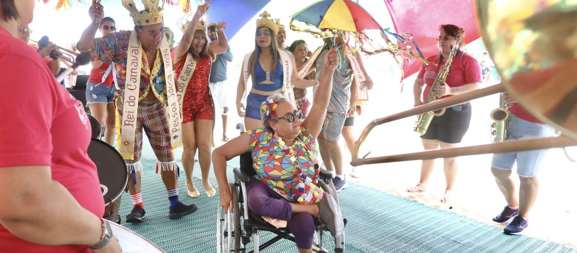 Fotografia de uma mulher parda, de cabelos grisalhos e óculos, sentada em uma cadeira de rodas enquanto segura uma sombrinha de frevo. Ao fundo, Luzia Santos, Rainha do Carnaval 2024, e Roberto Cabral, Rei do Carnaval 2024, acompanhados por outras pessoas fantasiadas e uma orquestra,
