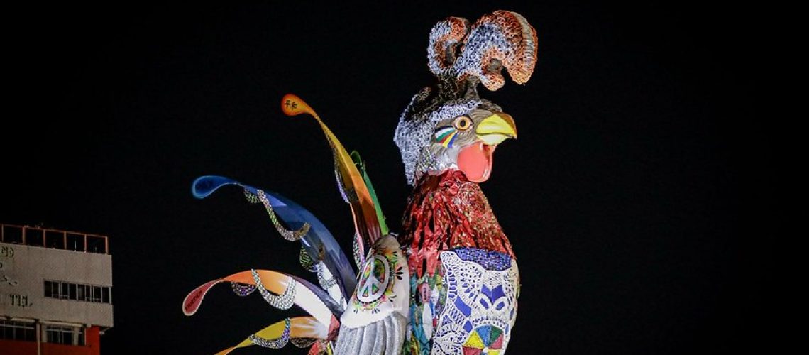 Galo da Madrugada iluminado à noite, decorado com mosaicos coloridos, rendas e detalhes vibrantes. Seu bico amarelo e a cauda exibe penas estilizadas em tons de azul, verde, vermelho e amarelo. No fundo, ceú todo preto.