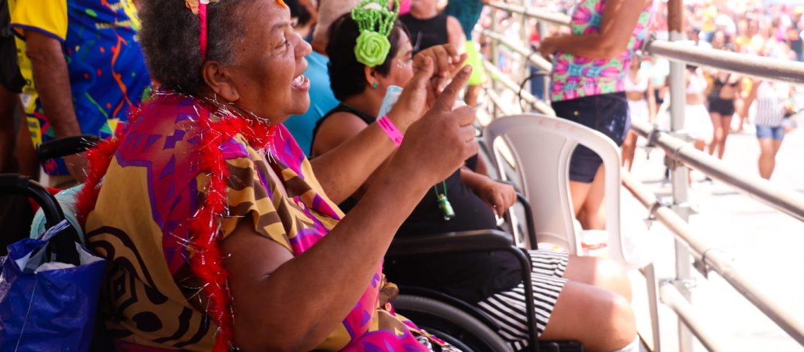 Fotografia de uma mulher parda, sorridente, vestindo roupas coloridas, sentada em uma cadeira de rodas de frente para uma grade. Ao fundo, várias pessoas trajando roupas carnavalescas estão sentadas.