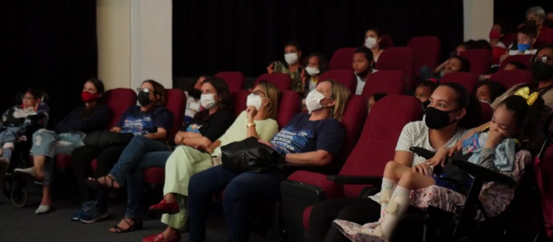 fotografia de uma sala de cinema com cadeiras vermelhas e várias pessoas espalhadas olhando para frente,