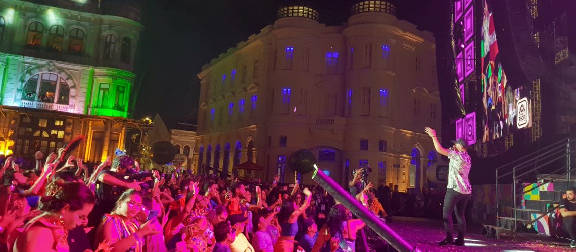 Um grupo de 25 pessoas com deficiência está reunido em frente ao palco. Atrás delas, uma plateia animada, com foliões em trajes carnavalescos. Ao fundo, edifícios históricos iluminados em tons de verde, azul e roxo compõem o cenário.