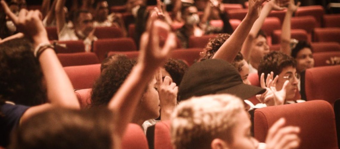 Fotografia de várias crianças sentadas no cinema com as mãos levantadas.